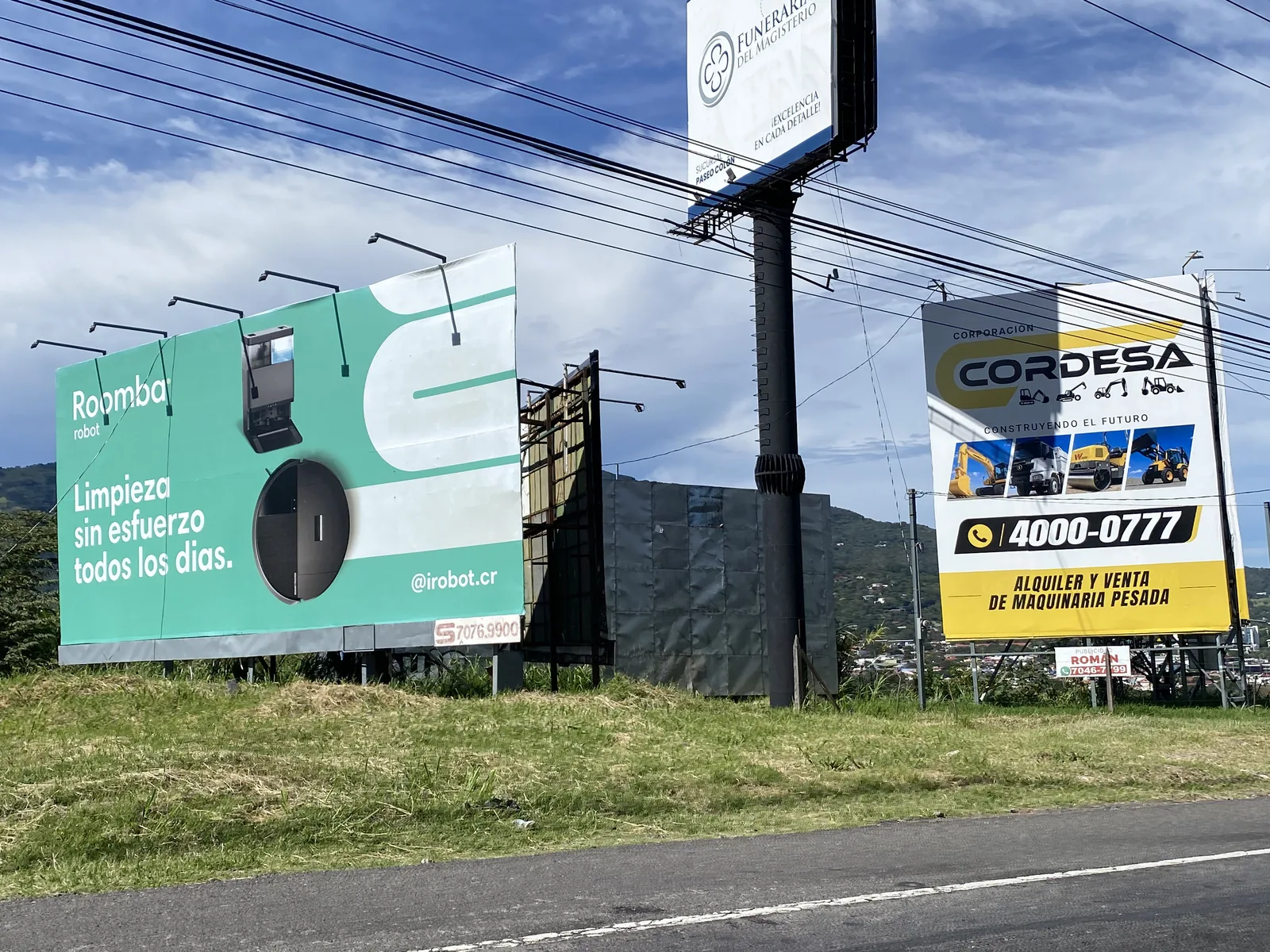 Vallas publicitarias de gran tamaño a la orilla de una carretera, con cables eléctricos encima, frente a los cerros.