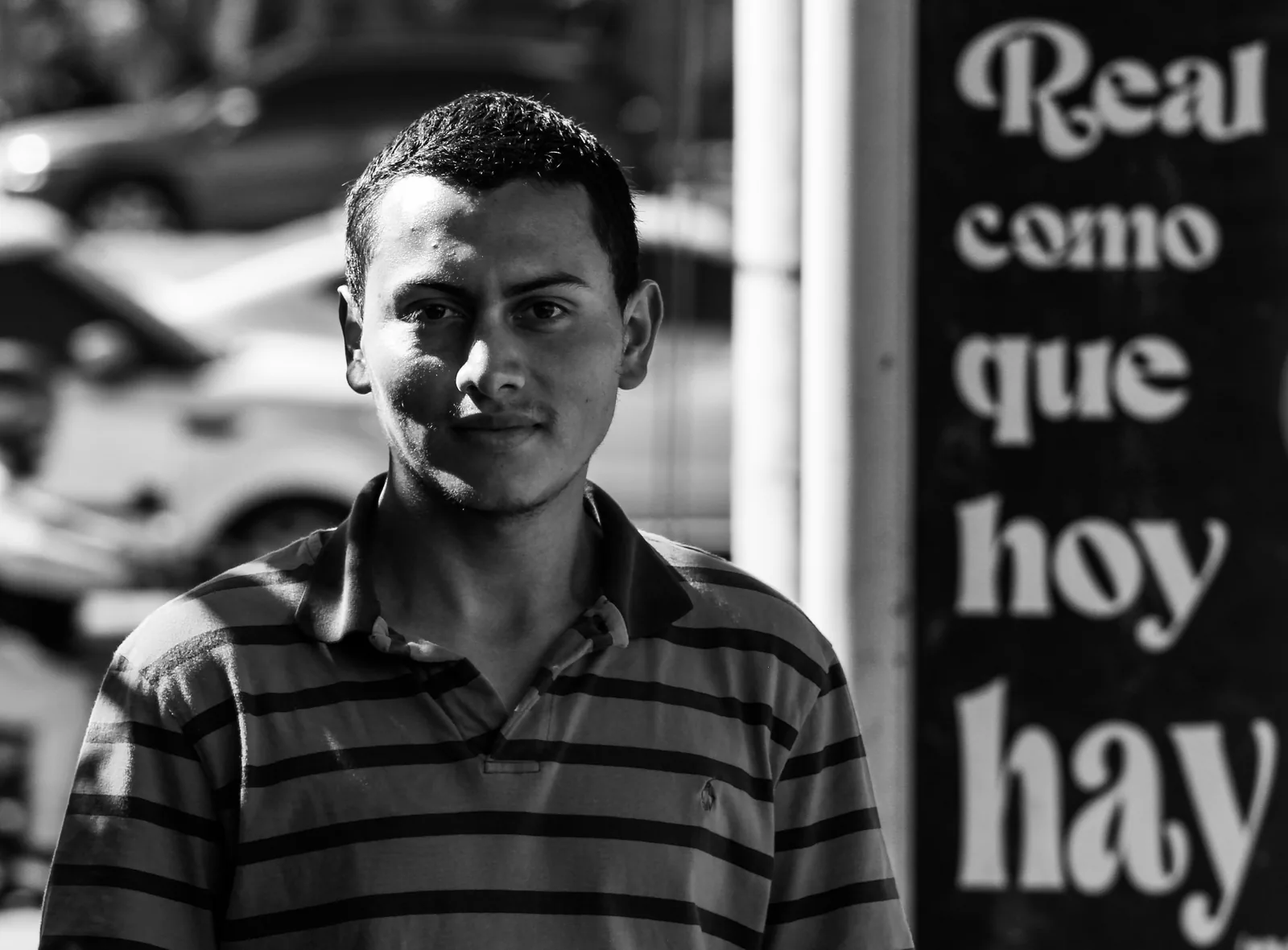 Retrato en blanco y negro de un joven con camisa a rayas; a su lado, un rótulo dice «Real como que hoy hay».