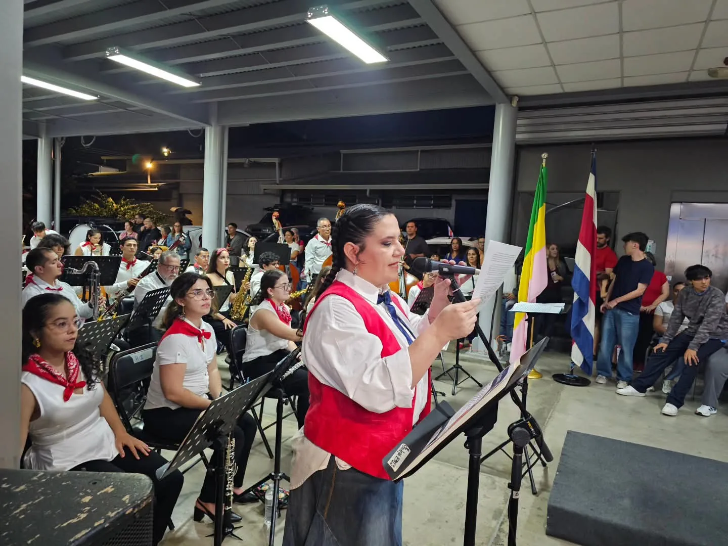 Una mujer con chaleco rojo lee un discurso frente a un micrófono; detrás, la banda de la EMAI, con camisa blanca y pañuelo rojo, y la bandera de Costa Rica.