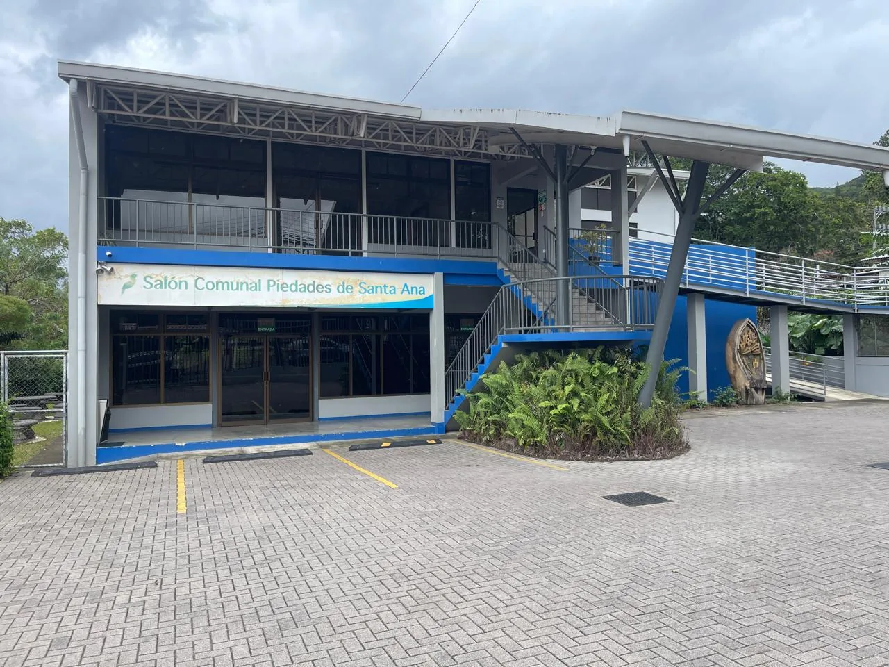 Fachada del Salón Comunal de Piedades de Santa Ana: un edificio de dos pisos con detalles azules y un parqueo adoquinado al frente.